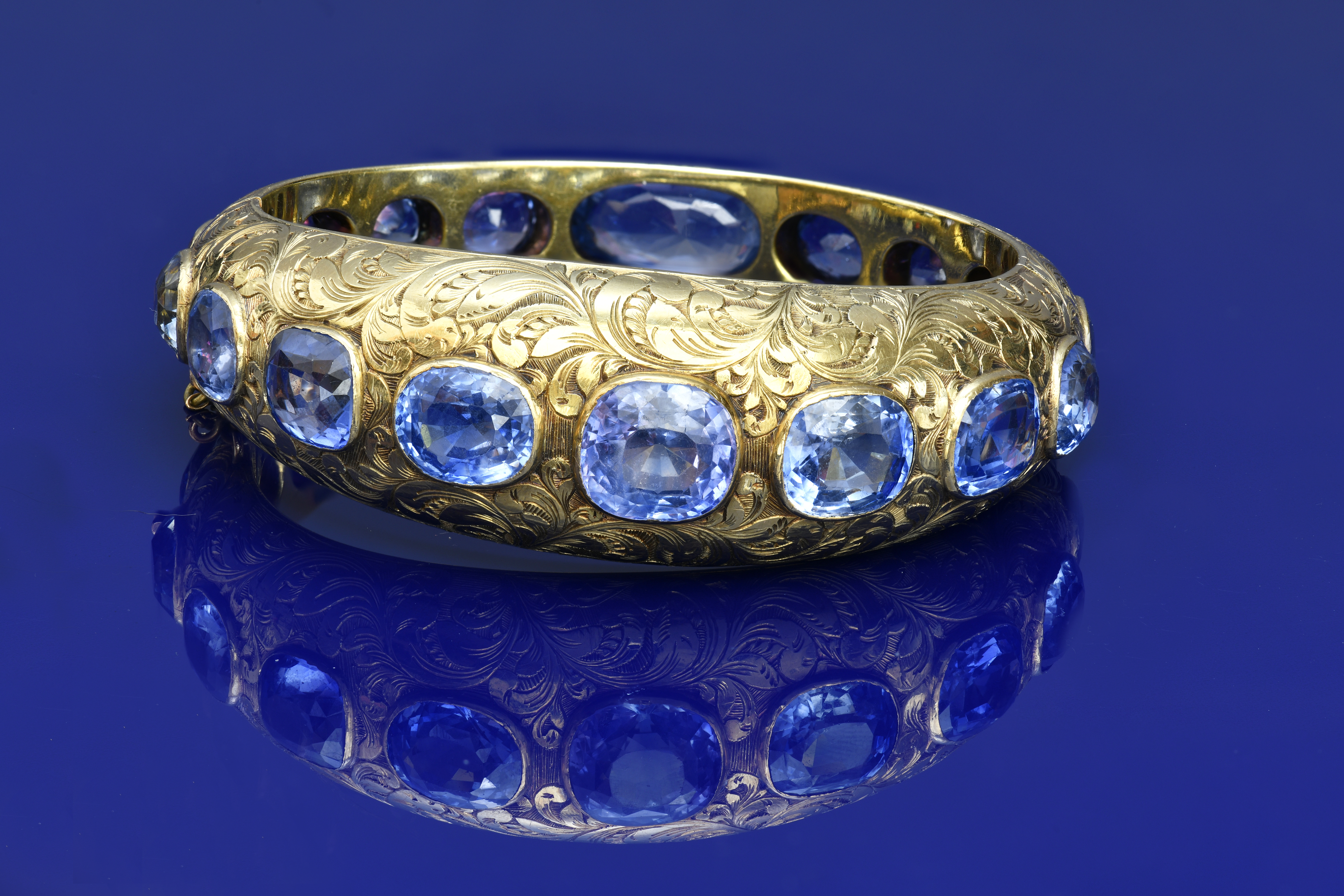 A c1900 18ct gold bangle with engraved floral decoration, set with seventeen cushion cut Ceylon Sapphires, the total sapphire weight approximately 68ct, 55.3g